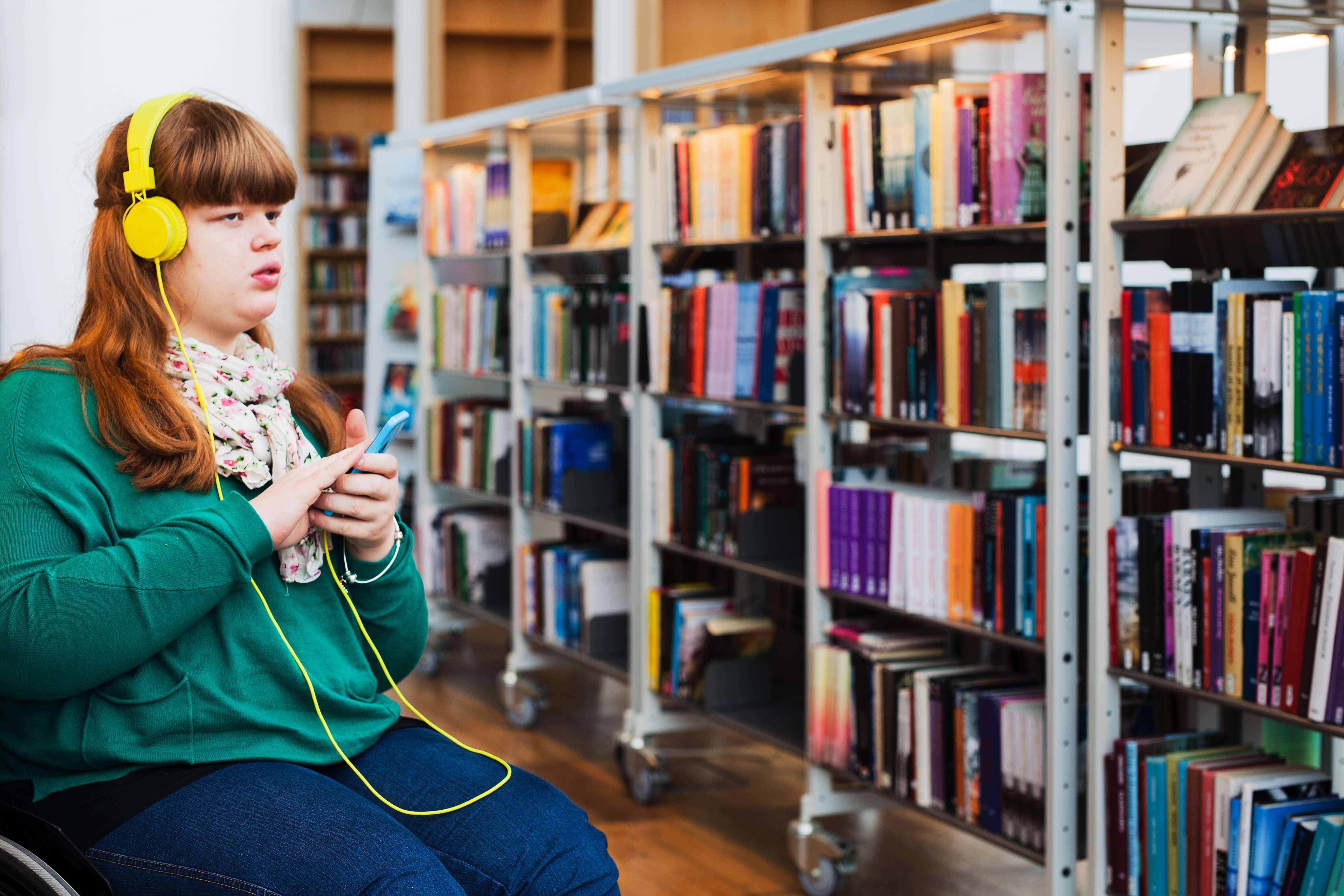 En ung kvinna sitter i en rullstol och lyssnar genom hörlurar på något i sin mobiltelefon. Omgivningen är ett bibliotek.